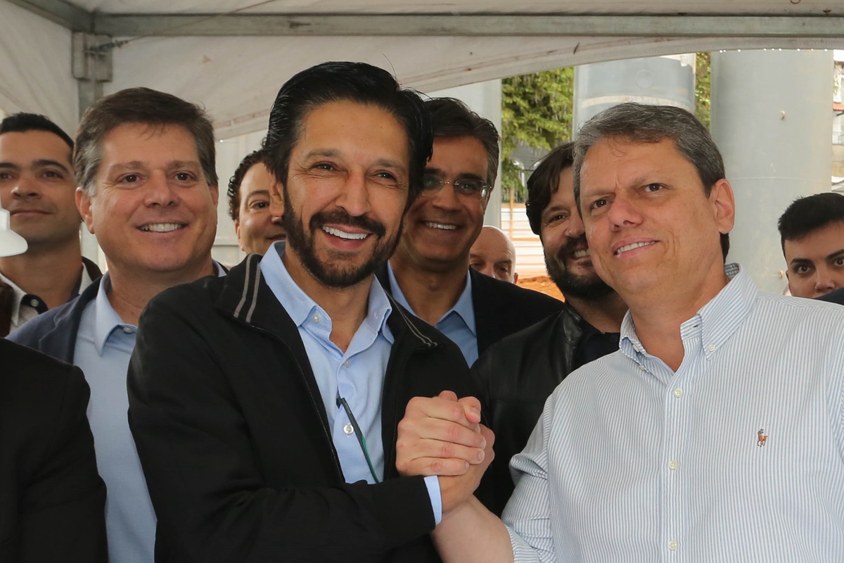 Imagem colorida mostra Ricardo Nunes, homem branco, de cabelo e barba pretos, camisa azul e blusa preta, de mãos dadas com Tarcísio de Freitas, homem branco, grisalho, de camisa branca