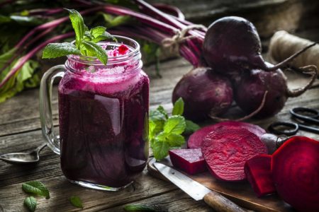 Bebida saudável: copo cheio de suco de beterraba orgânico fresco, tiro na mesa da cozinha de madeira rústica. Beterrabas inteiras e fatiadas estão espalhadas pelo copo. Uma faca de cozinha, uma colher e alguns ramos de hortelã completam a composição.