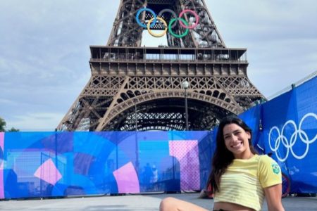 Imagem colorida mostra mulher branca com cabelos escuros longos e lisos. Ela veste camiseta amarela com listras brancas. Ao fundo está a Torre Eiffel com arcos olímpicos pendurados nela - Metrópoles