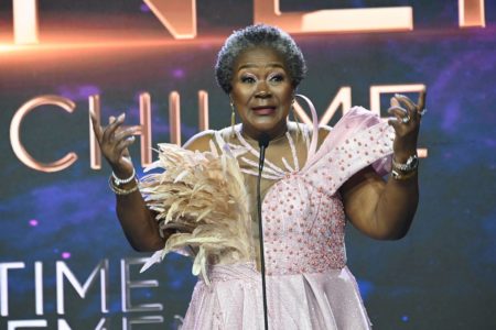 Na imagem colorida, Connie Chiume fala em microfone durante evento com vestido de festa claro - Metrópoles
