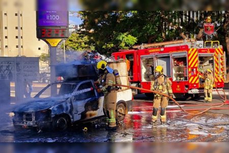 Imagem colorida de dois bombeiros apagando um incêndio em um carro branco. No fundo, há uma viatura vermelha do Corpo de Bombeiros do DF