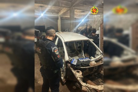 Imagem colorida de um carro sem a maioria das peças. À esquerda, um policial militar de farda azul observa o carro