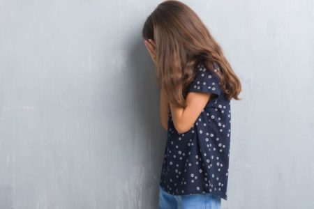 Foto colorida de menina, sobre parede cinza, com expressão triste, cobrindo o rosto com as mãos enquanto chora. Imagem ilustrativa - Metrópoles