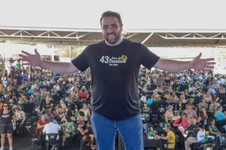 Homem branco , com ambos os braços abertos, sobre palco, com multidão ao fundo, em dia ensolarado - Metrópoles