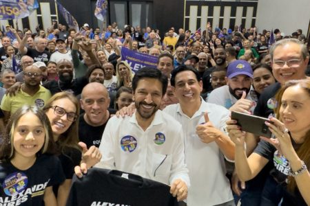 Imagem colorida mostra Ricardo Nunes, homem branco de cabelo e barba pretos,, vestidon camisas branca com adesivos eleitorais colados no peito, cercado de um auditório repleto de apoiadores - Metrópoles