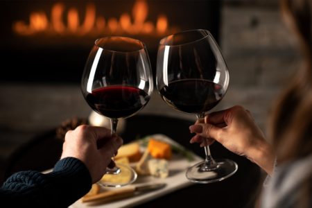 Close-up de mãos de casais torcendo por taças de vinho tinto em frente à lareira com tábua de queijos