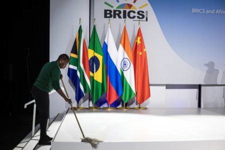 Imagem colorida mostra homem varrendo o chão, com bandeiras dos países do Brics ao fundo - Metrópoles