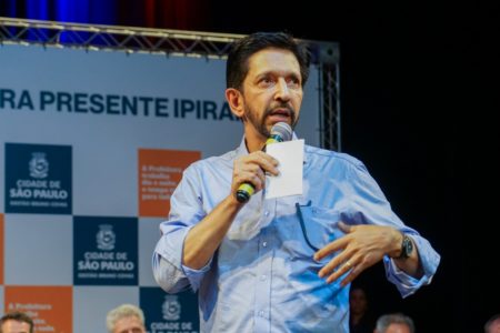 Imagem colorida mostra Ricardo Nunes, homem branco, de meia idade, de cabelo e barba pretos, de tipo magro, vestindo camisa azul clara de mangas arregaçadas, em imagem da cintura para cima. Ele está em um palco, falando ao microfone e olhando para o horizonte, com um banner da prefeitura de fundo - Metrópoles