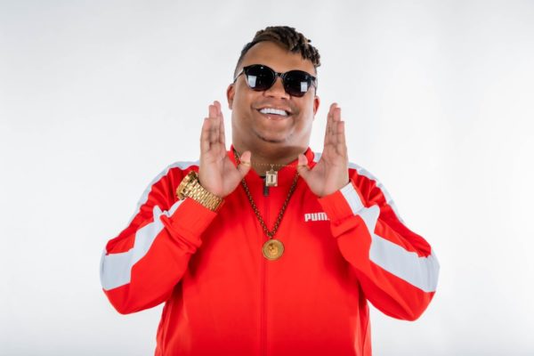 MC Delux posa, sorridente e de moletom vermelho, segurando o colar - Metrópoles