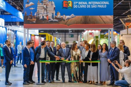 Imagem colorida mostra homem branco grisalho, vestindo terno. Ele está ao centro de diversos homens e mulheres e segura uma fita em cores verde e amarela - Metrópoles