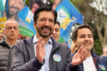 Imagem colorida mostra Ricardo Nunes, homem branco, de cabelo e barba tingidos de preto, em imagem do peito para cima, batendo palmas. ele veste jaqueta cinza e camisa azul clara. Está cercado de apoiadores políticos e bandeiras de campanha - Metrópoles