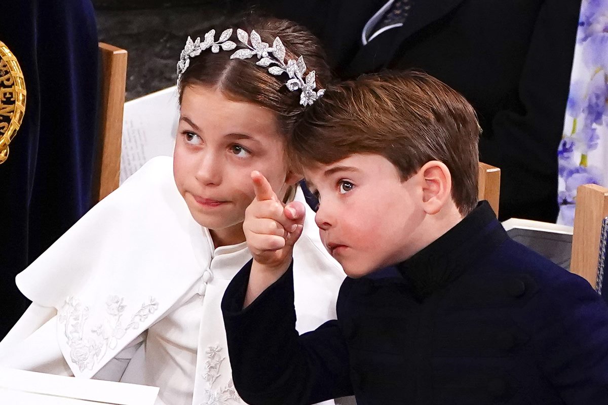 Foto colorida de menina com roupa branca e tiara na cabeça. Ela conversa com menino que aponta para algo. Ele usa uma roupa preta - Metrópoles