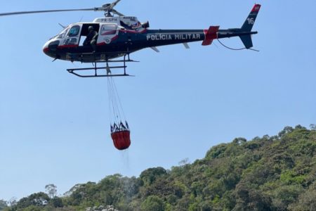 Imagem colorida mostra helicóptero sobrevoando área de mata carregando uma espécie de tonel com água - Metrópoles