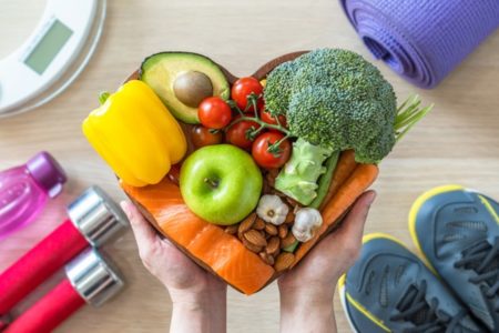 Pessoa segura prato em formato de coração com frutas e legumes. Balança, tênis, halteres e tapete de malhar estão ao redor - Metrópoles