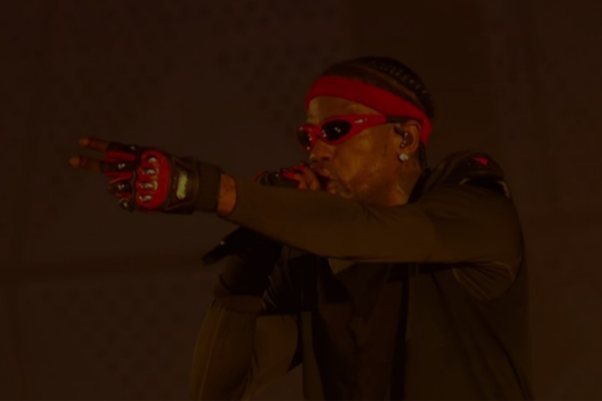 A imagem mostra o rapper Travis Scott durante um show. Com o microfone na mão direita, o artista canta usando bandana e óculos vermelho, junto a u par de luvas na mesma cor.