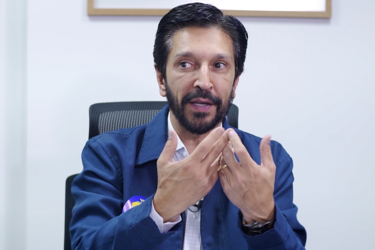 Imagem colorida mostra Ricardo Nunes, homem branco, de cabelo e barba pretos, vestindo jaqueta azul e camisa branca, sentado em uma cadeira com uma parede branca ao fundo, conversando - Metrópoles