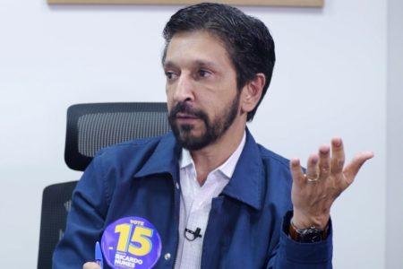 Imagem colorida mostra Ricardo Nunes, homem branco, de cabelo e barba pretos, vestindo jaqueta azul e camisa branca, sentado em uma cadeira com uma parede branca ao fundo, conversando - Metrópoles