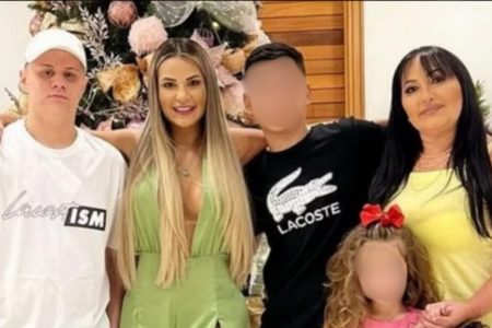 Foto de família caucasiana abraçada, da esquerda para direita: rapaz de boné e camiseta branca; mulher loira de cabelos longos soltos, abaixo dos seios, com vestido verde musgo; rapaz de camiseta preta, com desenho de jacaré branco, abaixo dele, menina de cabelo encaracolado, loiro, com laço vermelho; ao lado dela, mulher de meia idade de camiseta amarela e cabelos longos pretos - Metrópoles