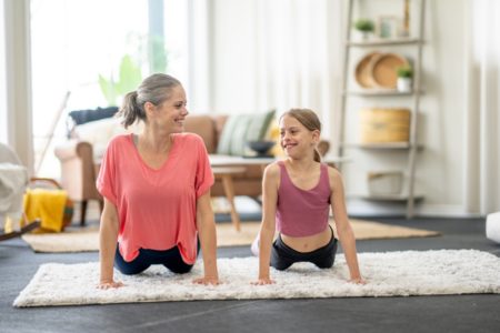 Mulher de cabelos grisalhos e menina se olham enquanto praticam yoga em sala - Metrópoles - Testes