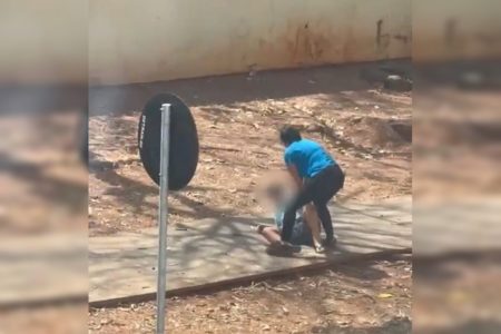 Imagem colorida de uma mulher, de camiseta azul e calça preta, segurando o filho, caído em uma calçada rodeada de terra