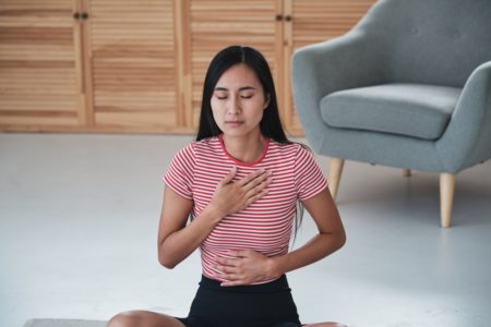 Mulher asiática praticando ioga e exercícios respiratórios pela manhã, sentada em posição de lótus com os olhos fechados, segurando as mãos no peito e estômago, meditando em casa