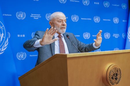 Imagem colorida de fundo azul com símbolo da ONU e Lula à frente discursando - Metrópoles