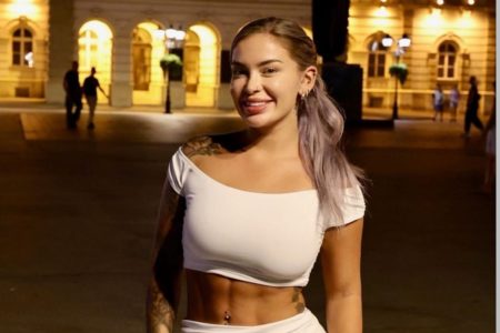 Foto de Hayley Davies. Uma mulher branca, loira, usando um cropped branco e sorrindo para a foto. Metrópoles