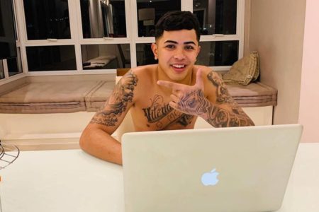 Imagem colorida de MC Vini VL sentado em cadeira, fazendo sinal com a mão e em frente de computador - Metrópoles