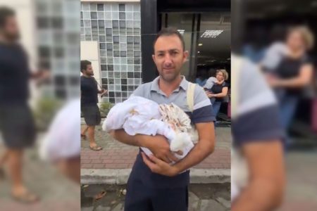 Imagem colorida mostra homem branco segurando no colo bebê enrolado em cobertas - Metrópoles