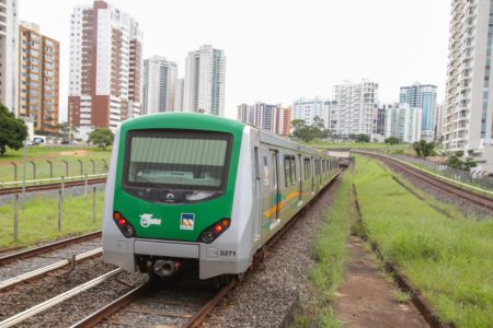 Imagem colorida de um trem verde com cinza sobre os trilhos. Ao redor, grama verde