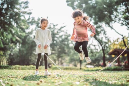 Foto colorida de duas meninas brincando de pular corda em área verde - Metrópoles