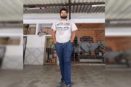 Imagem colorida de um homem negro em pé, com as mãos no bolso. Ele veste camiseta branca e calça jeans azul