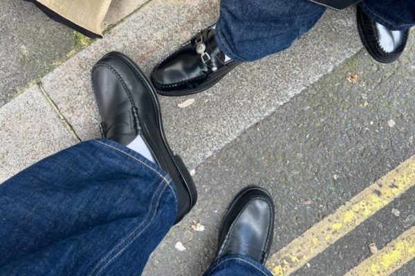 A imagem mostra dois pares de sapatos pretos pisando no asfalto