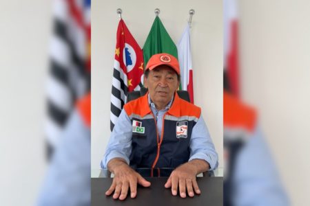 Imagens coloridas mostram homem sentado à mesa, de colete azul e laranja, boné laranja, sentado atrás de mesa, ao fundo bandeiras de São Paulo (à esquerda), do Brasil (centro) e de Taboão da Serra (à direita)