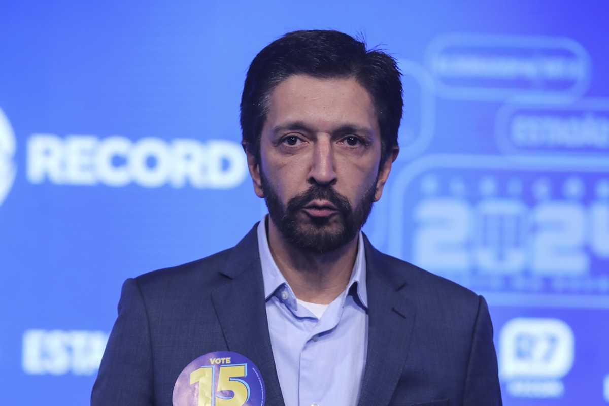Imagem colorida mostra Ricardo Nunes, homem branco de cabelo curto e barba pretos, de terno azul escuro e camisa azul clara, falando com um telão azul ao fundo - Metrópoles