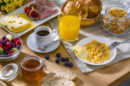 Foto colorida de mesa com café da manhã, com suco, ovos mexidos, presunto, mel e frutas - Metrópoles
