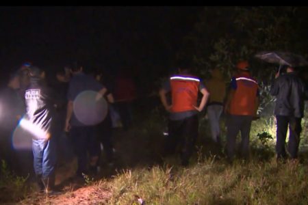 Imagem colorida mostra pessoas de costas, em área de mata. Alguns homens vestem coletes alaranjados com faixa reflexora. Está escuro ao redor - Metrópoles