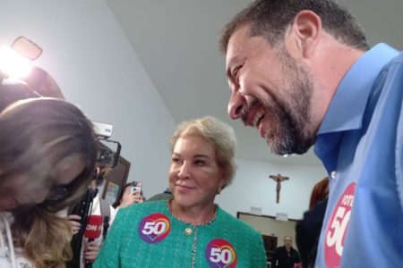 Imagem colorida mostra, no meio, mulher loira de cabelos curtos. Ela veste casaco verde claro com dois adesivos com o número 50. No seu lado esquerdo está um homem branco com cabelos escuros e barba fina, do outro lado aparece cabelo castanho claro de uma mulher - Metrópoles