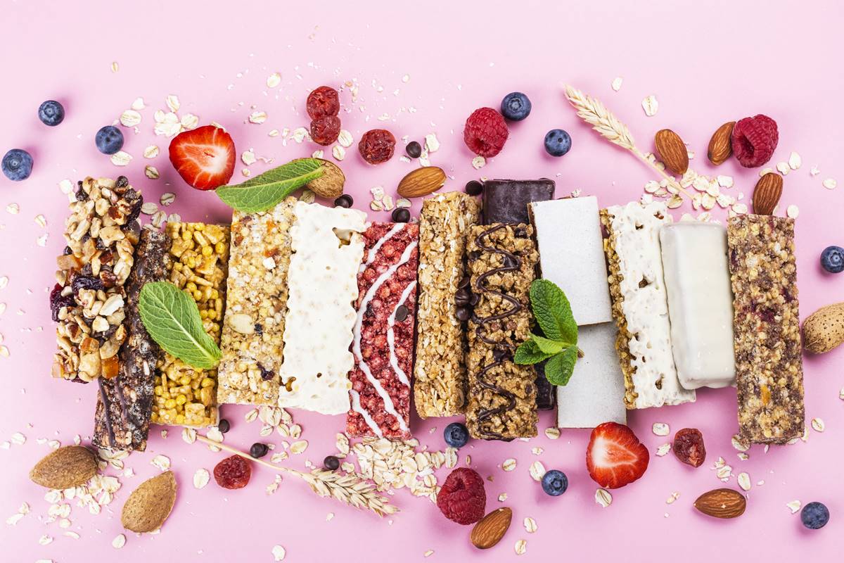 Foto colorida de barras de cereais e de proteína em fundo rosa. Há frutas e granola ao redor dos alimentos - Metrópoles