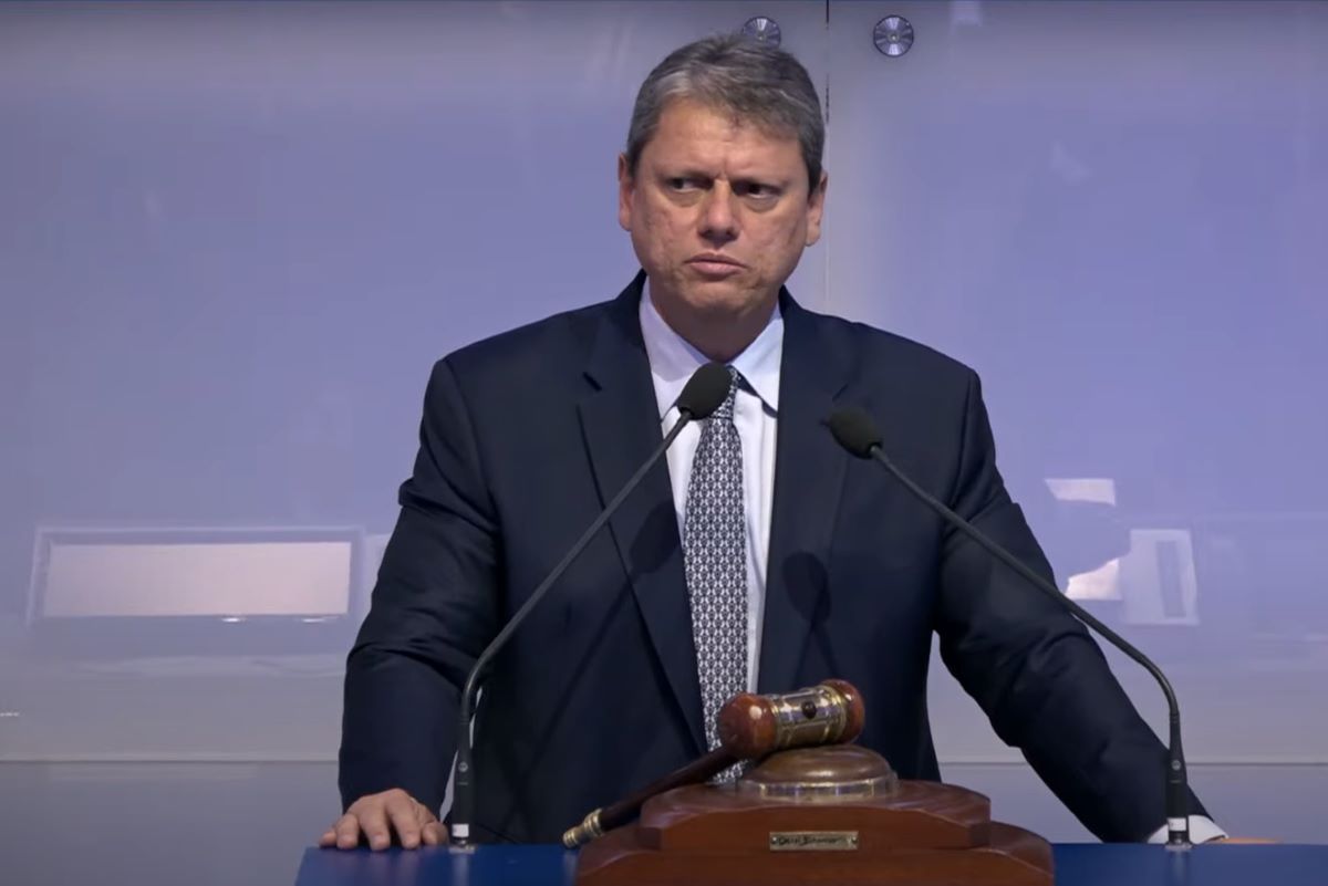 imagem colorida mostra governador tarcísio de freitas. ele veste um terno preto, com gravata cinza. está atrás de dois microfones em um púlpito que também contém um martelo