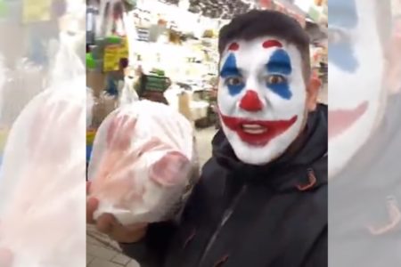 Imagem colorida de um homem com filtro de palhaço no rosto segurando uma cabeça de porco embalada em plástico - Metrópoles