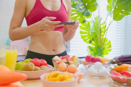Foto colorida de mulher com roupa de malhar perto de uma mesa com frutas e legumes. Ela está tirando fotos - Metrópoles