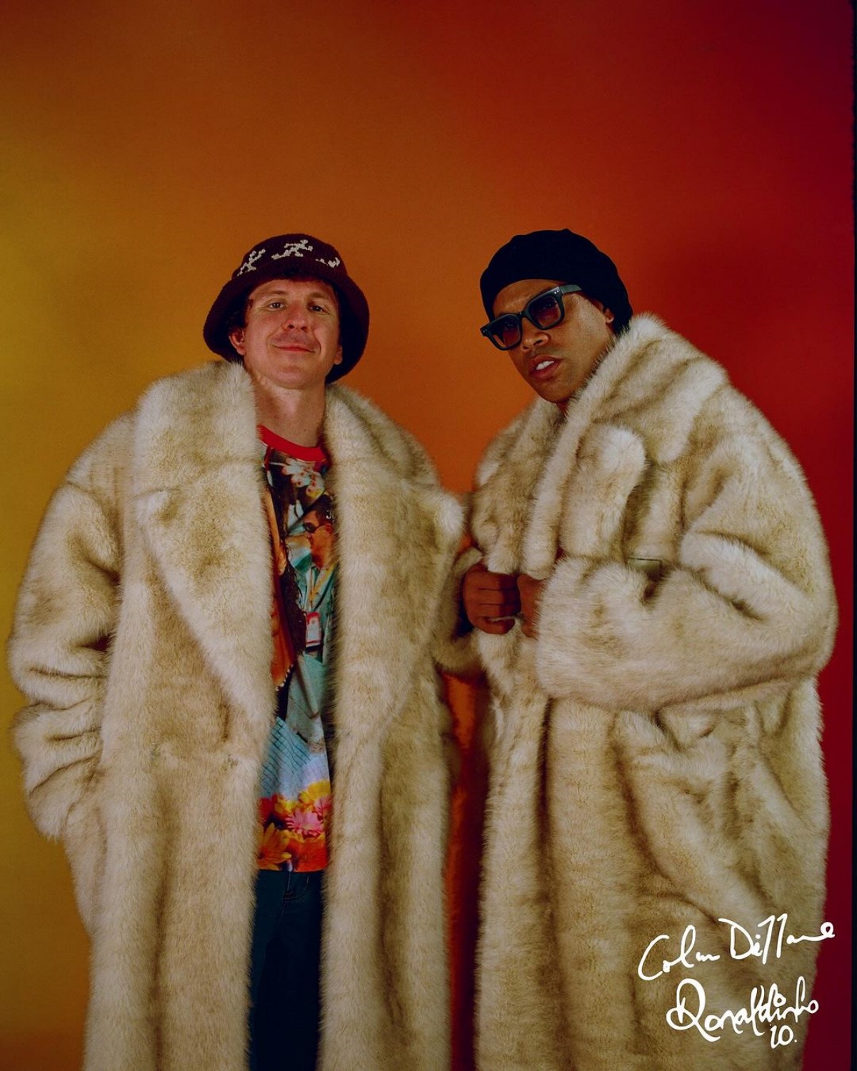 A imagem mostra Ronaldinho e KidSuper posando para a foto. Ambos olham para a câmera enquanto vestem o mesmo modelo de casaco sobretudo peludo.