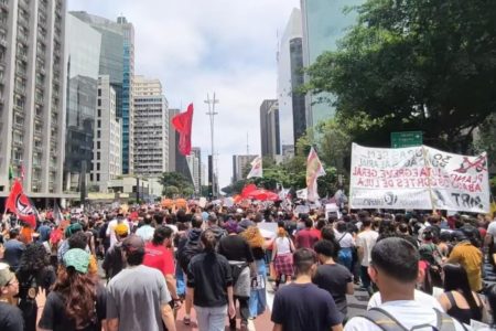 Imagem colorida mostra aglomeração de pessoas segurando faixas vermelhas em avenida - Metrópoles