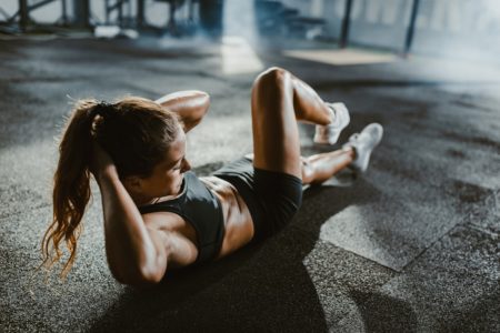 Jovem atleta exercitando abdominais com as mãos atrás da cabeça em uma academia. Metrópoles