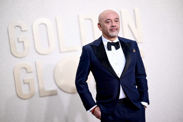 A imagem mostra o designer Christian Louboutin vestindo um smoking preto com camisa branca em um fundo branco