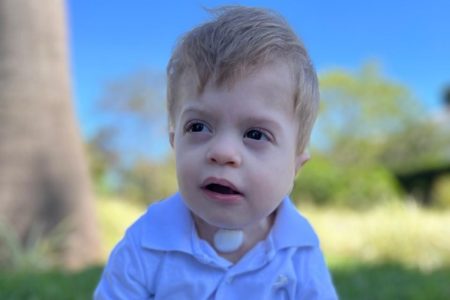 Foto colorida de menino loiro de blusa azul sentado em grama - Metrópoles