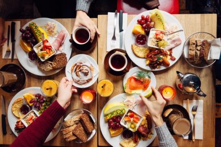 Imagem mostra uma mesa cheia de pratos e comidas vista de cima. Vários praços brindam com taças - Metrópoles