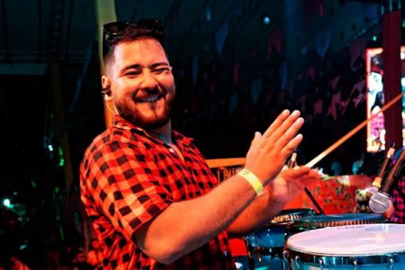 Imagem colorida de homem sorrindo, usando blusa xadrez e tocando percussão - Metrópoles