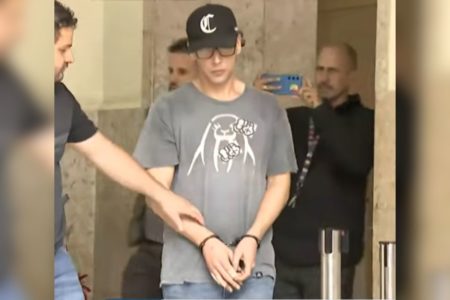 Imagem colorida de homem branco de porte magro. Ele tem as mãos à frente do corpo, presas com algemas. Veste calça jeans, camiseta cinza, boné preto e óculos. Ele tem o braço segurado por um homem com colete da polícia - Metrópoles